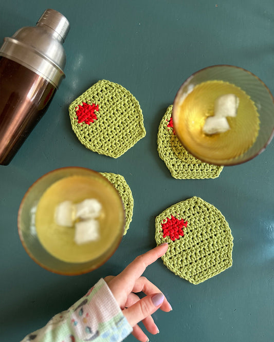 A hand sets a devouthand Ready-to-order ✧ Set of Olive coasters (hand-crocheted and green) on the table. Two glasses filled with green liquid and ice sit on matching coasters, with a cocktail shaker visible in the background.
