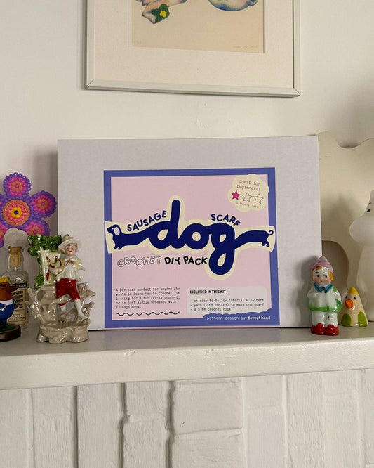 A "Sausage Dog scarf ✧ DIY pack" by devouthand is displayed on a white shelf, surrounded by decorative figurines and a flower ornament. A small tag suggests it's ideal for beginners.