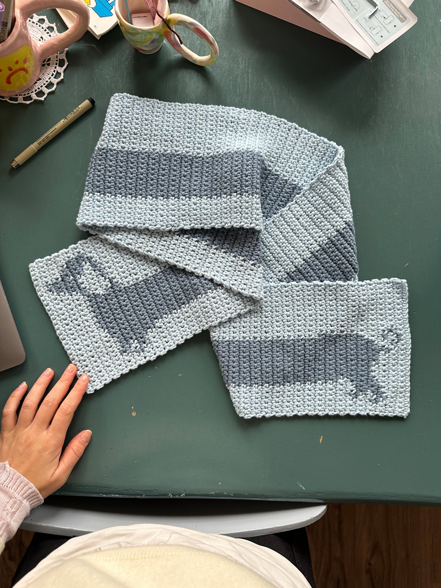 A hand touches a knitted devouthand Ready-to-order ✧ Handmade Sausage Dog scarf with wide blue stripes, draped over a dark green table. Nearby are a pen, decorative mug with brushes, and an open organizer.