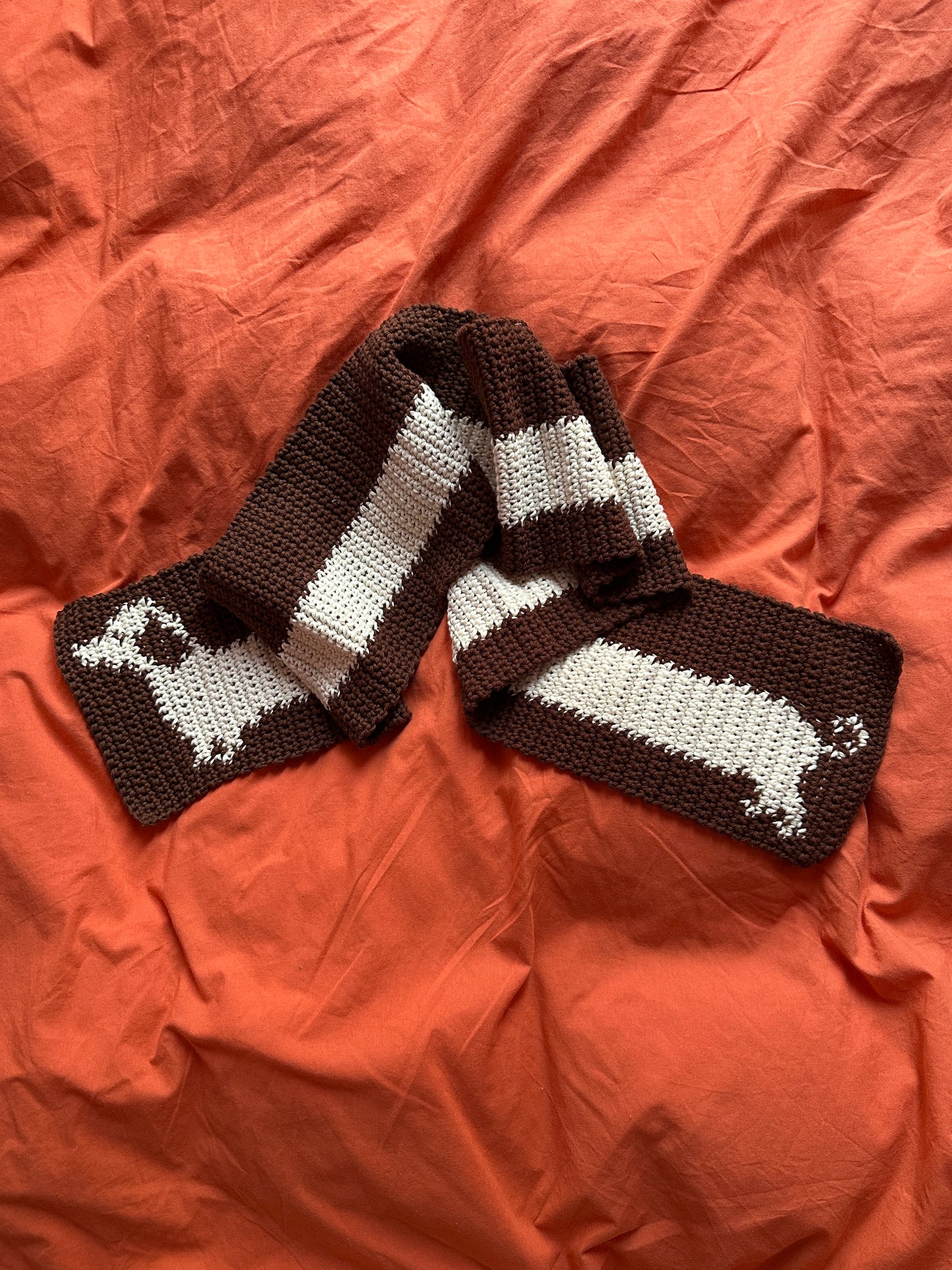 A hand touches a knitted devouthand Ready-to-order ✧ Handmade Sausage Dog scarf with wide blue stripes, draped over a dark green table. Nearby are a pen, decorative mug with brushes, and an open organizer.