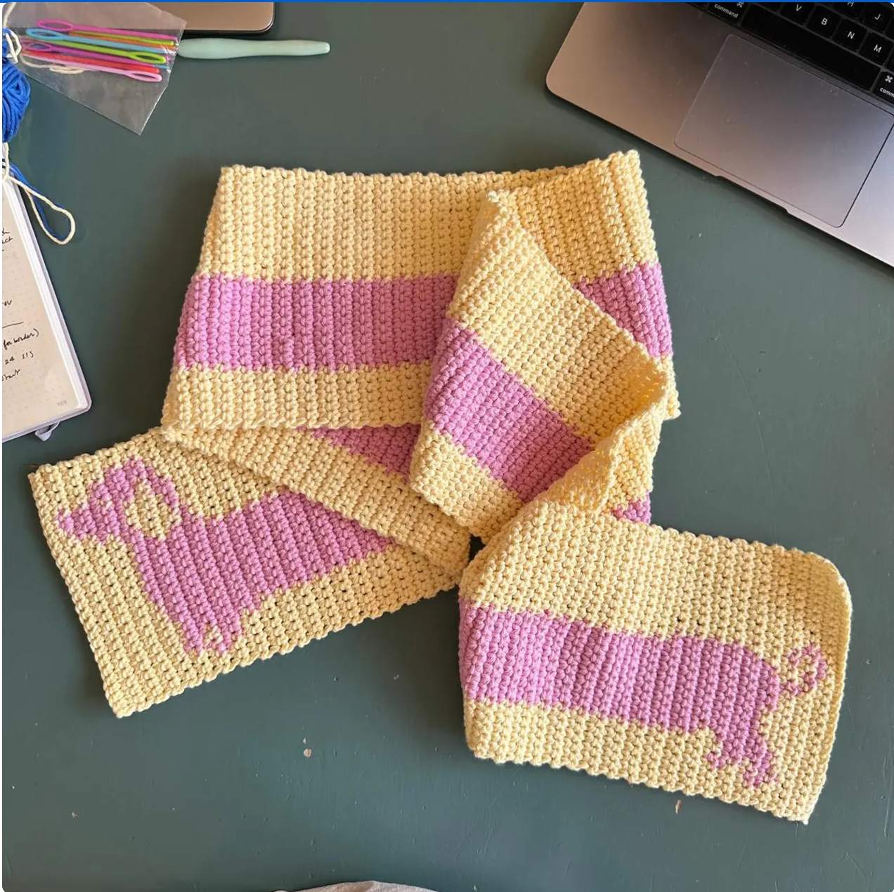 A hand touches a knitted devouthand Ready-to-order ✧ Handmade Sausage Dog scarf with wide blue stripes, draped over a dark green table. Nearby are a pen, decorative mug with brushes, and an open organizer.