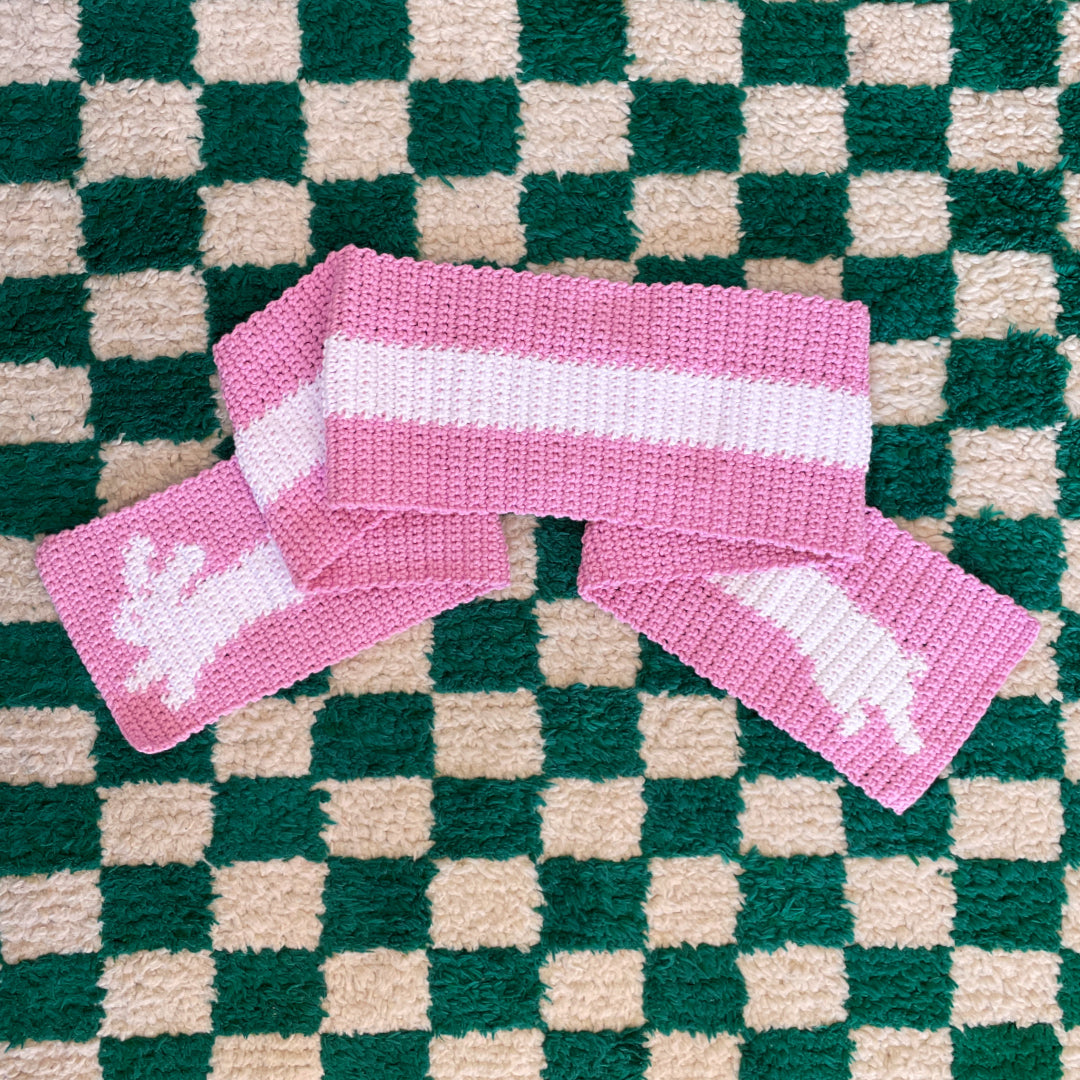 A stylish pink and white scarf from the "Crochet pattern combo pack ✧ Pet scarves" by devout hand, featuring a charming reindeer pattern on both ends, is displayed on a checkered green and white rug.