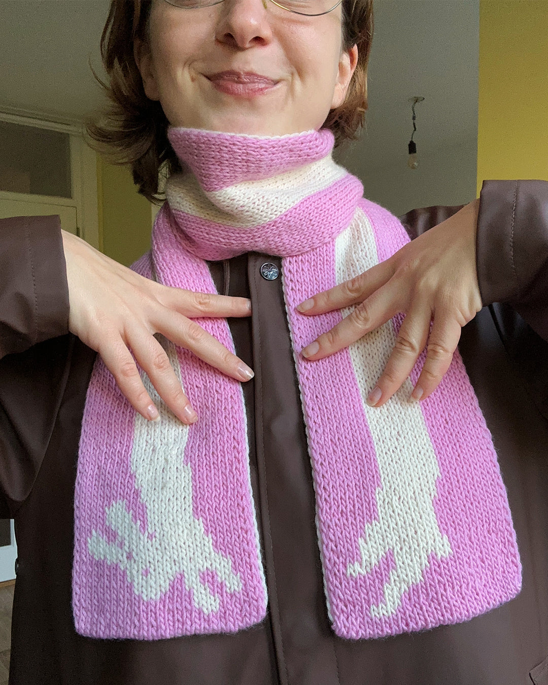 The devouthand Ready-to-order ✧ Bunny scarf, a pink and white double-knit featuring a giraffe design on one end, is displayed on a green and white checkered surface with a brown dog partially visible in the corner.
