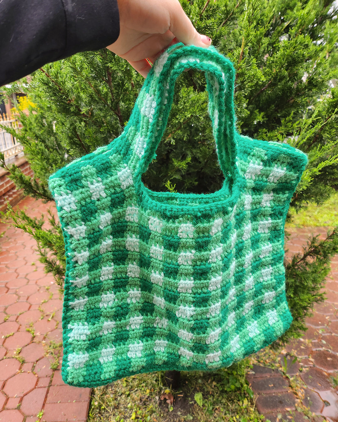 A picnic scene on a grassy lawn features the "Crochet bag pattern ✧ Picnic bag" by devouthand, showcasing its red and white checkered design on a blanket with a strawberry print. Nearby are wooden toy food items, an open illustrated book, and a person's knee visible in the bottom left.