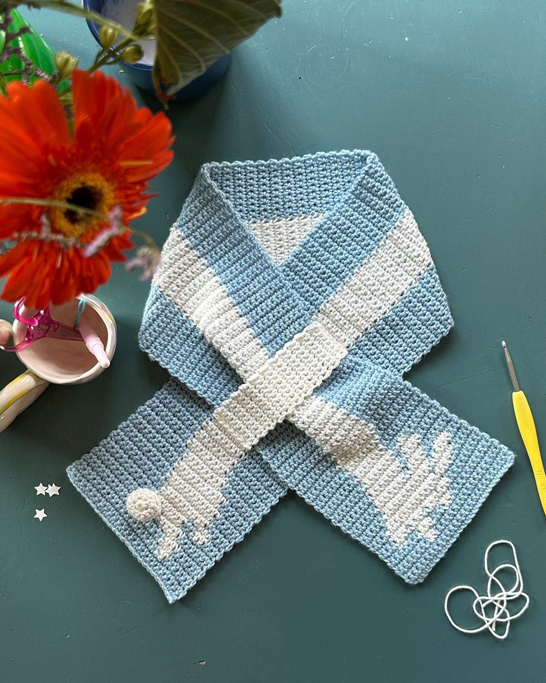 A person with glasses takes a mirror selfie in a well-lit room. They are wearing a white long-sleeve shirt, blue jeans, and the "Crochet scarf pattern ✧ Bunny (adult/kids size)" by devouthand in blue and yellow. Their hair is tied up, and they hold the phone in their right hand. The background features an open space.