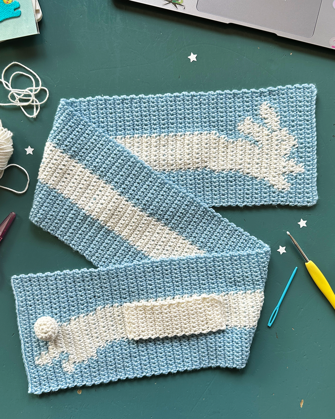 A person with glasses takes a mirror selfie in a well-lit room. They are wearing a white long-sleeve shirt, blue jeans, and the "Crochet scarf pattern ✧ Bunny (adult/kids size)" by devouthand in blue and yellow. Their hair is tied up, and they hold the phone in their right hand. The background features an open space.