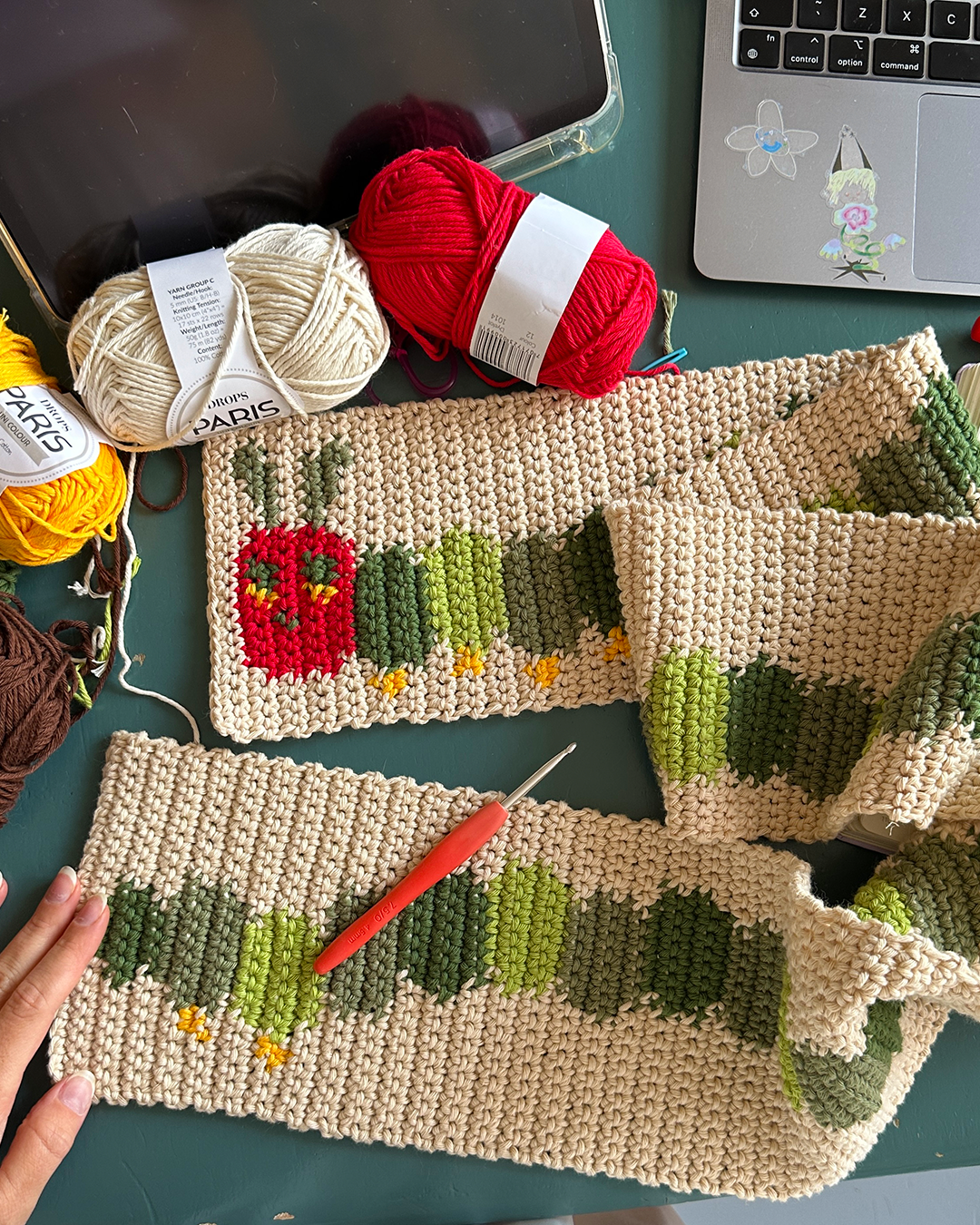 A crocheted scarf from devout hand, following the "Crochet scarf pattern ✧ Very Hungry Caterpillar (adult/kids size)," lies on a green and white checkered carpet. The caterpillar design features shades of green with a red head that includes eyes and antennae.