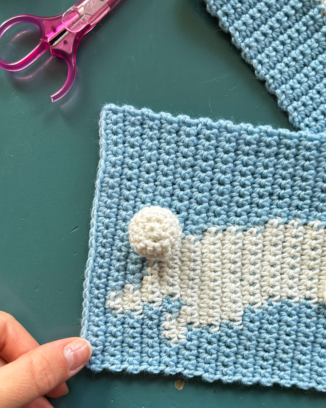 A person with glasses takes a mirror selfie in a well-lit room. They are wearing a white long-sleeve shirt, blue jeans, and the "Crochet scarf pattern ✧ Bunny (adult/kids size)" by devouthand in blue and yellow. Their hair is tied up, and they hold the phone in their right hand. The background features an open space.