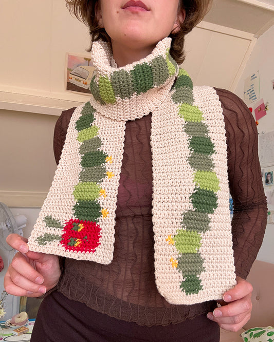 A crocheted scarf from devout hand, following the "Crochet scarf pattern ✧ Very Hungry Caterpillar (adult/kids size)," lies on a green and white checkered carpet. The caterpillar design features shades of green with a red head that includes eyes and antennae.