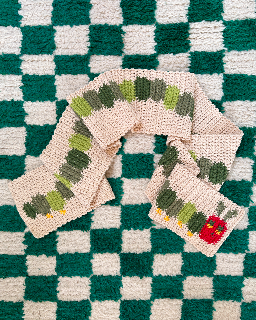 A crocheted scarf from devout hand, following the "Crochet scarf pattern ✧ Very Hungry Caterpillar (adult/kids size)," lies on a green and white checkered carpet. The caterpillar design features shades of green with a red head that includes eyes and antennae.